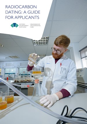 A person wearing a white lab coat, gloves and protective glasses, pouring a liquid into a beaker