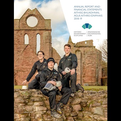 Three people wearing protective gear hold traditional tools and sit on a wall in front of a ruined abbey.
