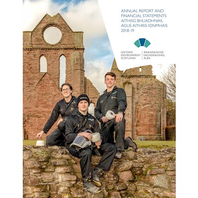 Three people wearing protective gear hold traditional tools and sit on a wall in front of a ruined abbey.