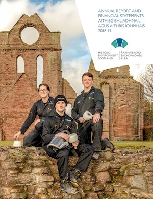 Three people wearing protective gear hold traditional tools and sit on a wall in front of a ruined abbey.