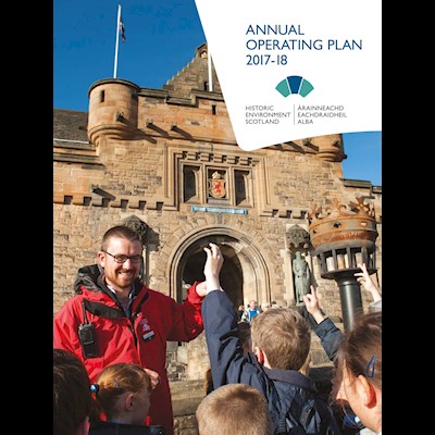 A cover of a publication with a man in a red jacket smiling and talking to a group of children in front of a castle on a sunny day