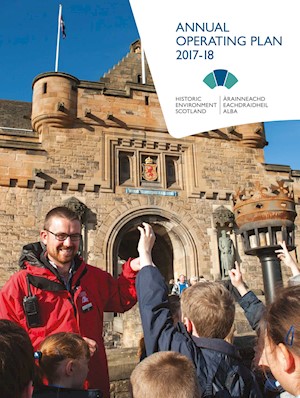 A cover of a publication with a man in a red jacket smiling and talking to a group of children in front of a castle on a sunny day